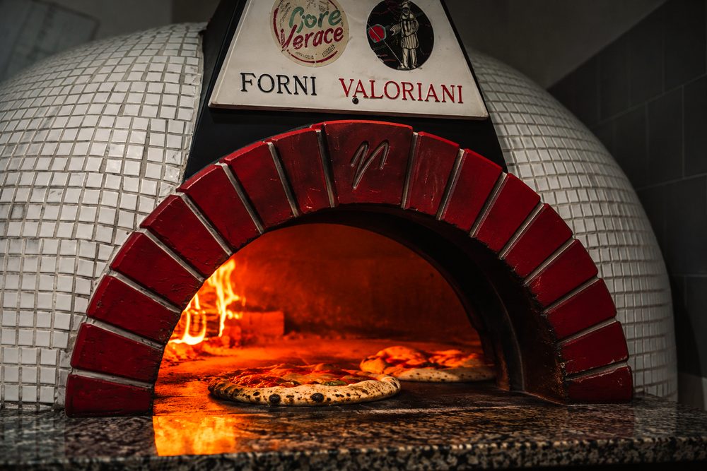 La bocca del forno Valoriani con la fiamma viva e due pizze in cottura sotto l'arco di mattoni rossi