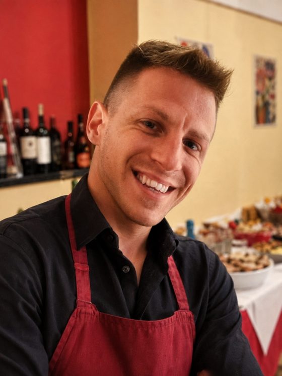 Sorriso in sala, col grembiule bordeaux e il muro delle bottiglie alle spalle