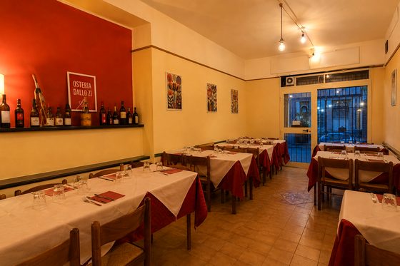 La sala con l'insegna Osteria dallo Zì sul muro rosso e la mensola delle bottiglie